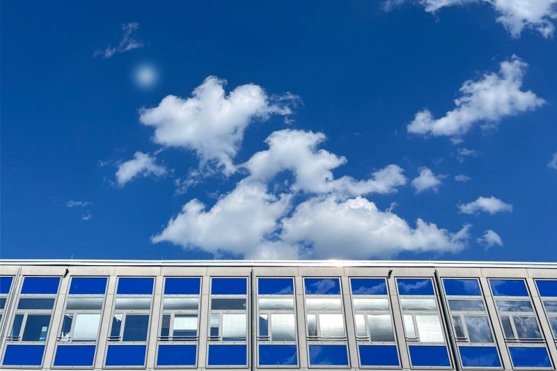 Moderne Gebäudefassade mit reflektierenden Fenstern vor klarem Blaumann und Wolken.