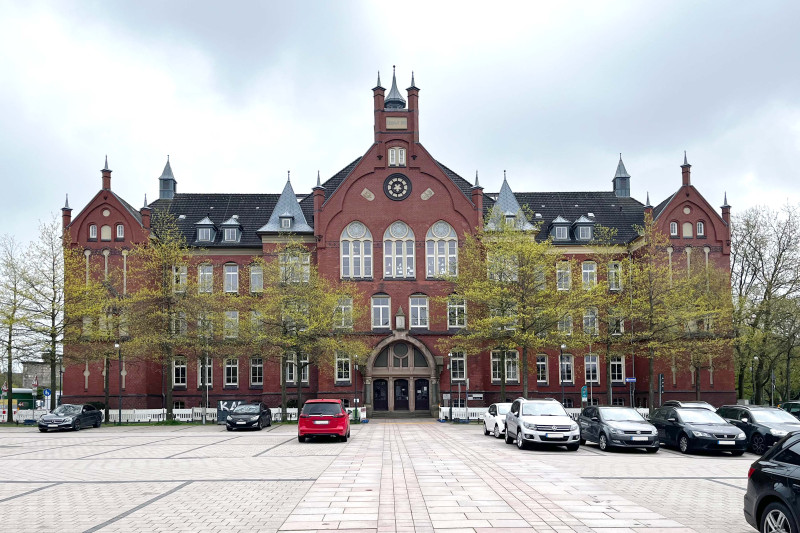 Historisches, rotes Gebäude mit Türmen, umgeben von Bäumen und parkenden Autos.