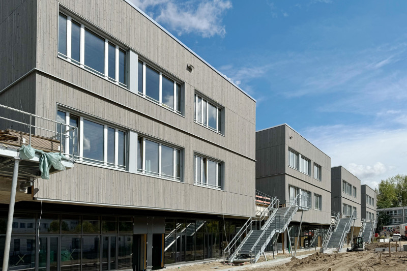 Moderne Neubauten mit Holzfassade und Treppen, umgeben von Baustellenatmosphäre und blauen Himmel.