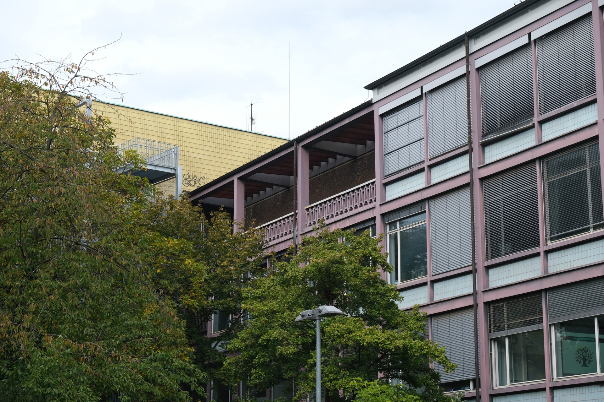 Ansicht eines modernen Gebäudes mit Bäumen und Balkon im Vordergrund, teils bewölkter Himmel.