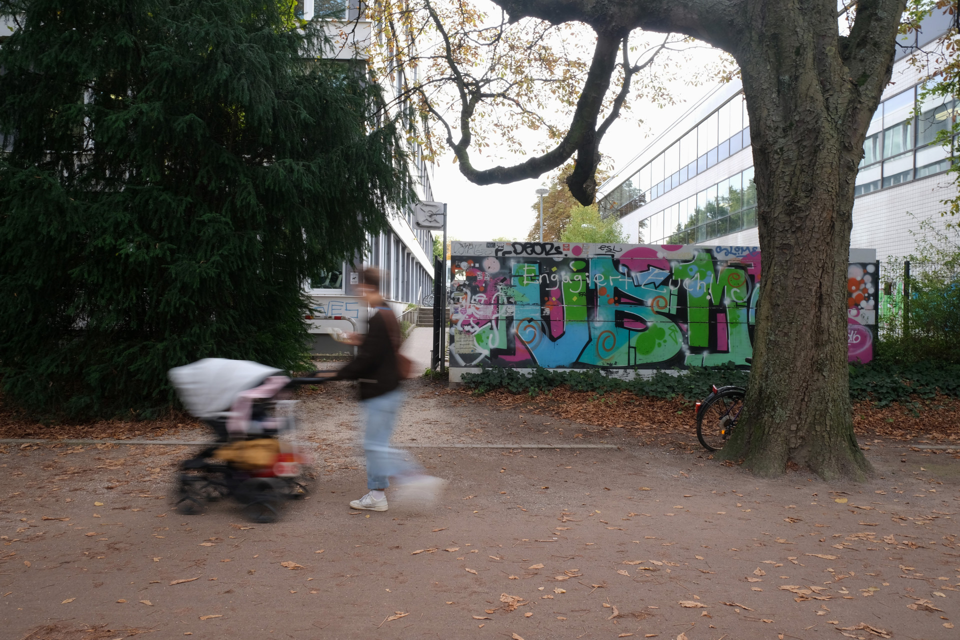 Eine Person mit Kinderwagen geht an einer bunten Graffiti-Wand vorbei, umgeben von Bäumen.