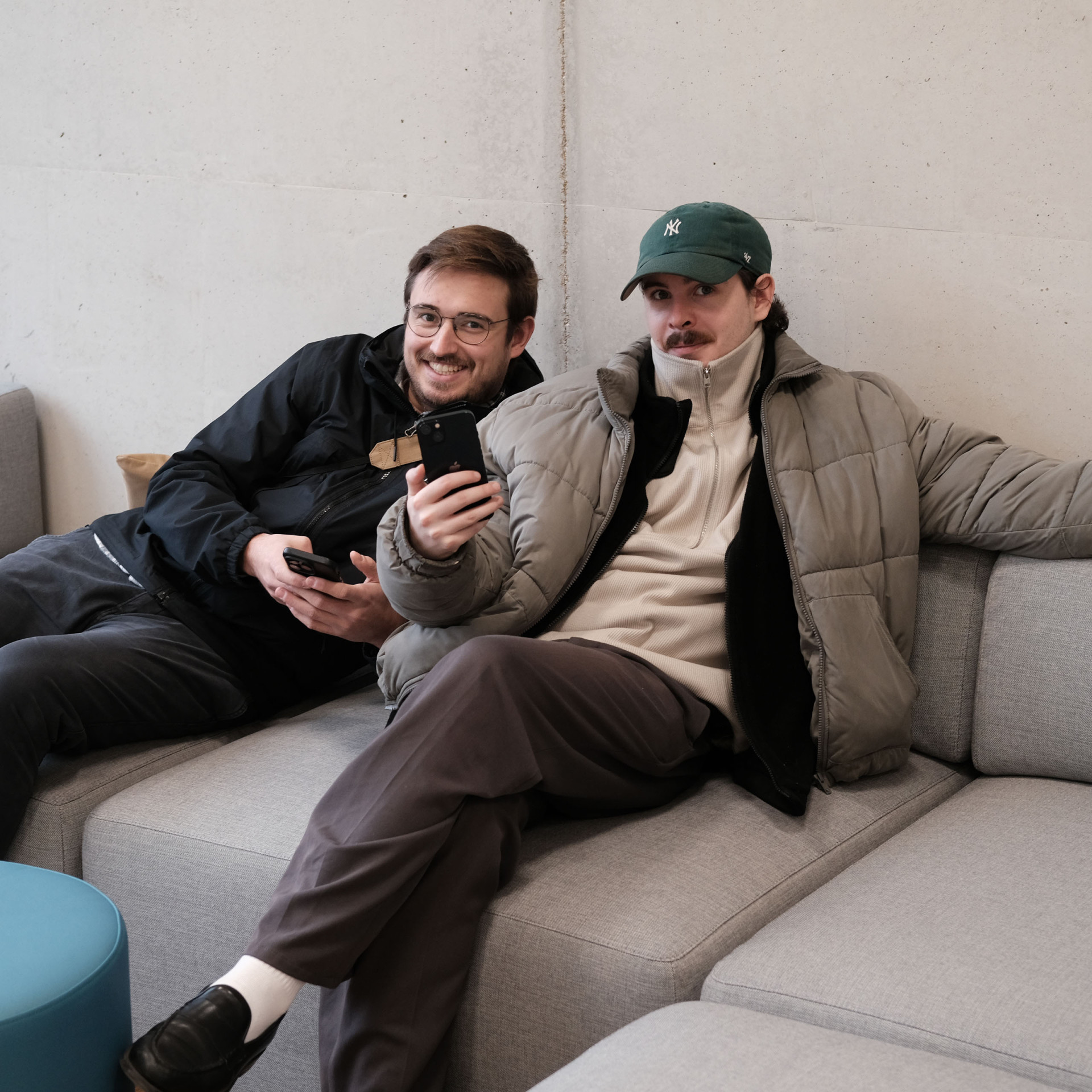 Zwei Männer sitzen entspannt auf einer grauen Couch und schauen auf ihr Smartphone.