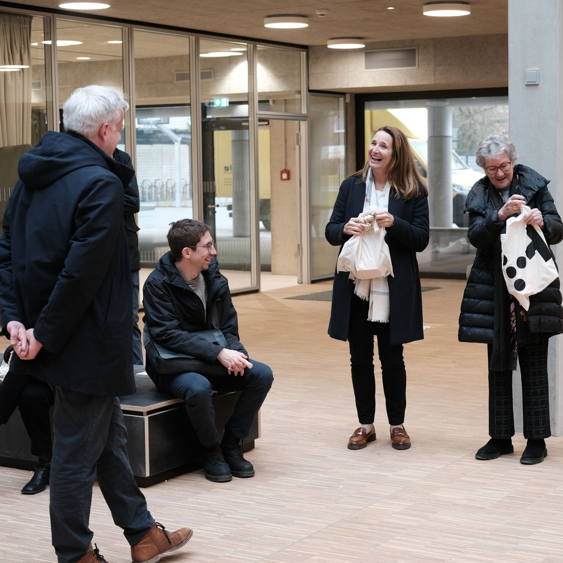 Gruppengespräch in modernem Raum, lachende Frauen mit Taschen, Männer hören gespannt zu.
