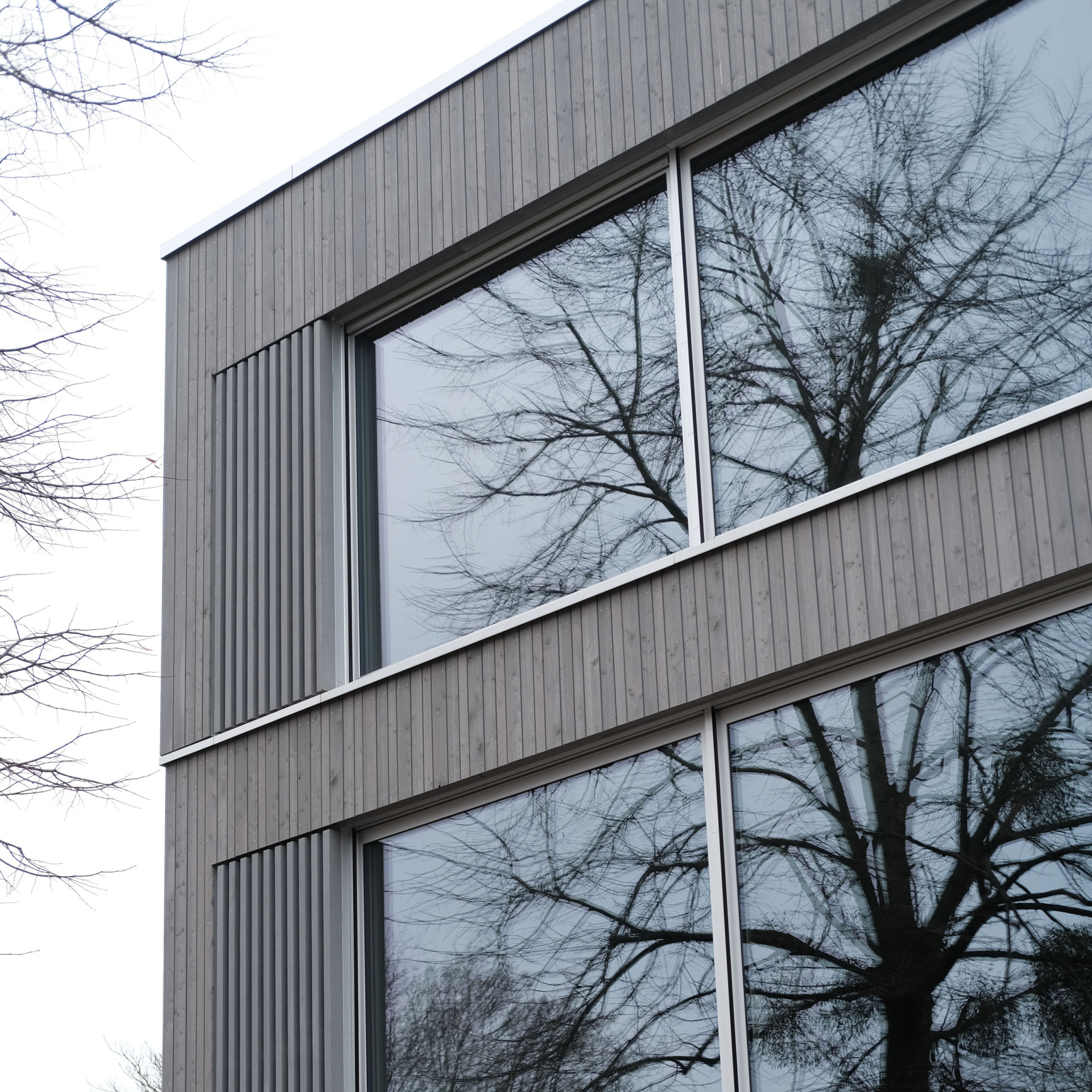 Modernes Gebäude mit großen Fenstern, die Bäume reflektieren, im grauen Licht des Tages.