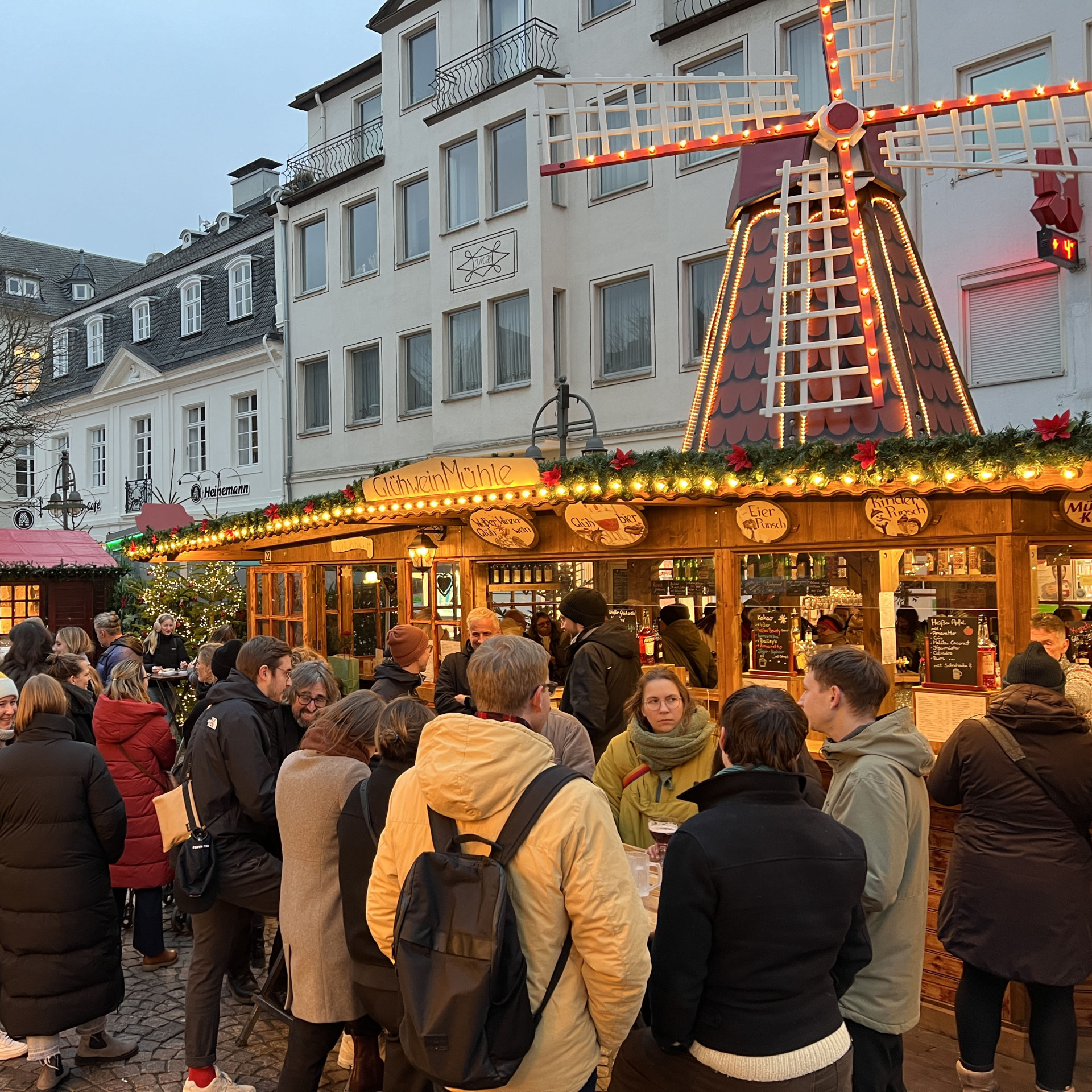 Beleuchteter Weihnachtsmarkt mit Menschenmenge vor einem Holzstand und einer Windmühle.