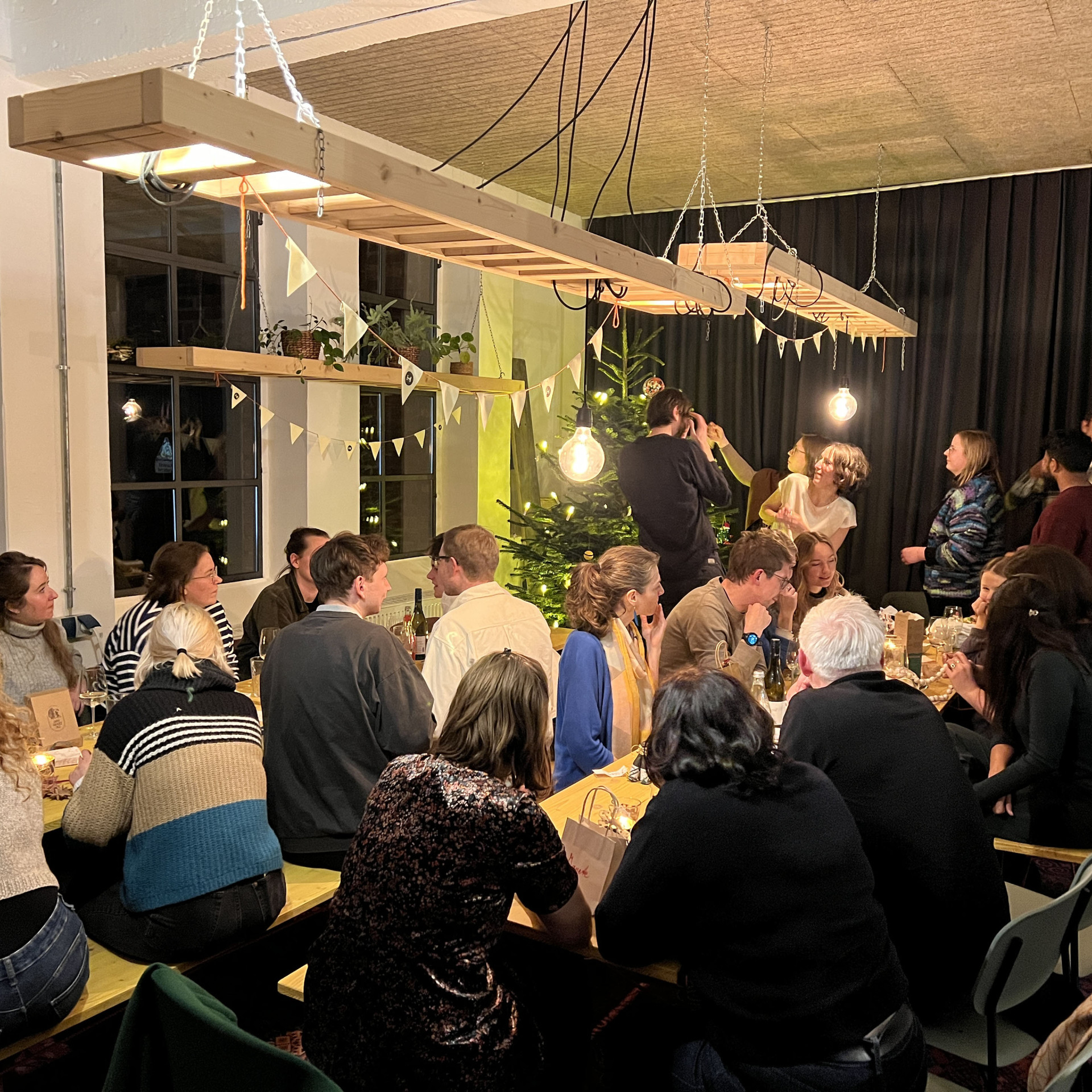 Gemütliches Festessen mit mehreren Gästen, Lichtern und Weihnachtsbaum im Hintergrund.