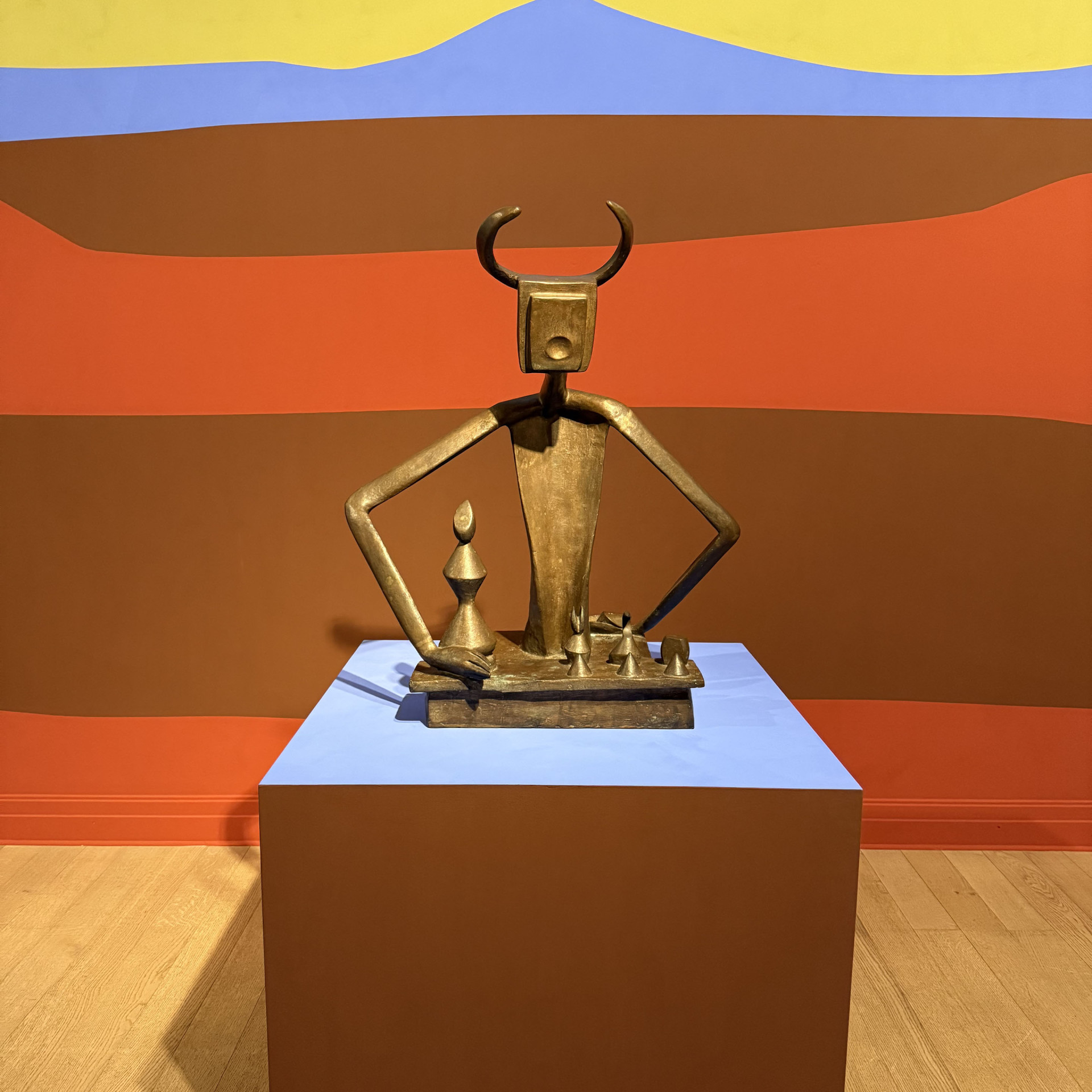 Bronzeskulptur mit Hörnern steht auf blauem Sockel, umgeben von buntem Wandhintergrund.