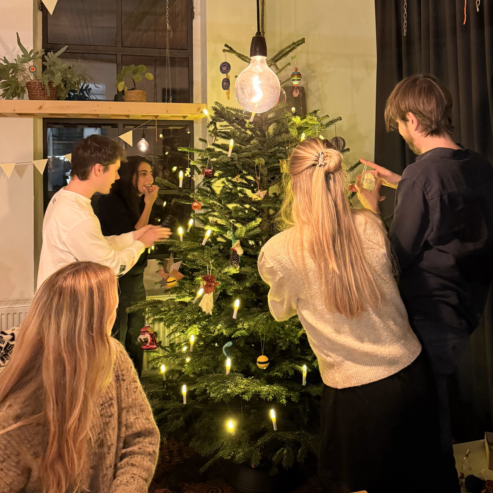 Freunde schmücken gemeinsam einen Weihnachtsbaum mit Lichtern und buntem Schmuck.