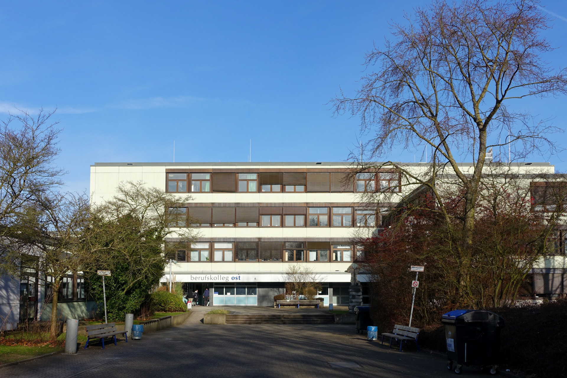 Modernes Berufskolleg ost mit klaren Linien, umgeben von Bäumen und blühenden Sträuchern.