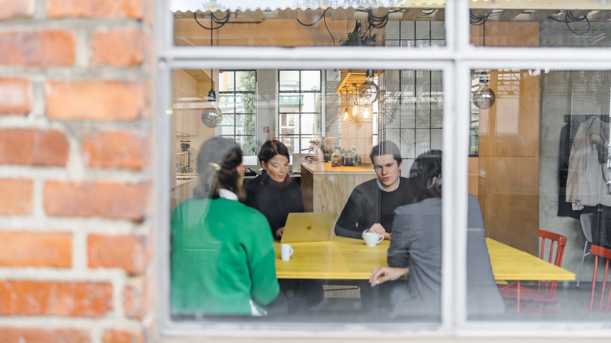 Außenperspektive zeigt den Blick in den Pausenraum, wo vier Mitarbeitende zusammen an einem gelben Tisch sitzen.