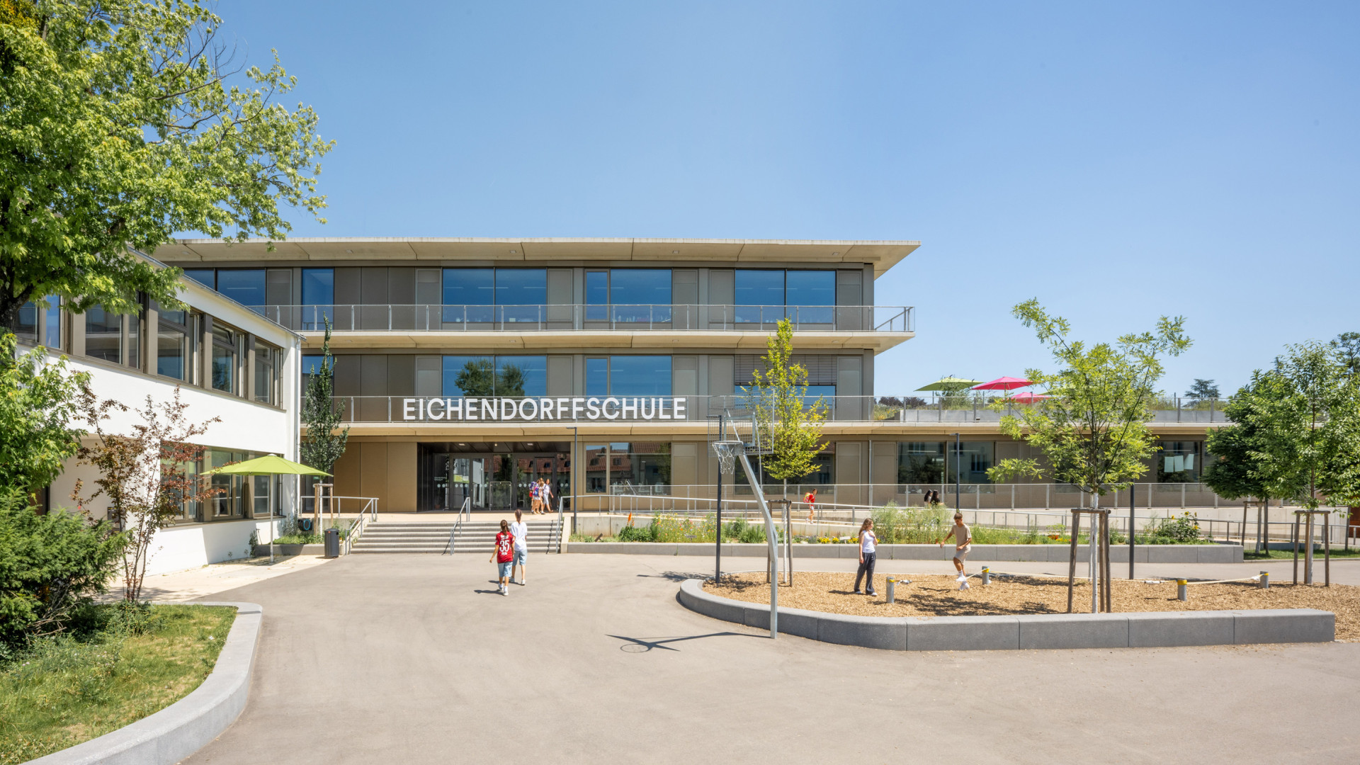 Moderne Eichendorffschule mit flacher Fassade, umgeben von Bäumen und spielenden Kindern.