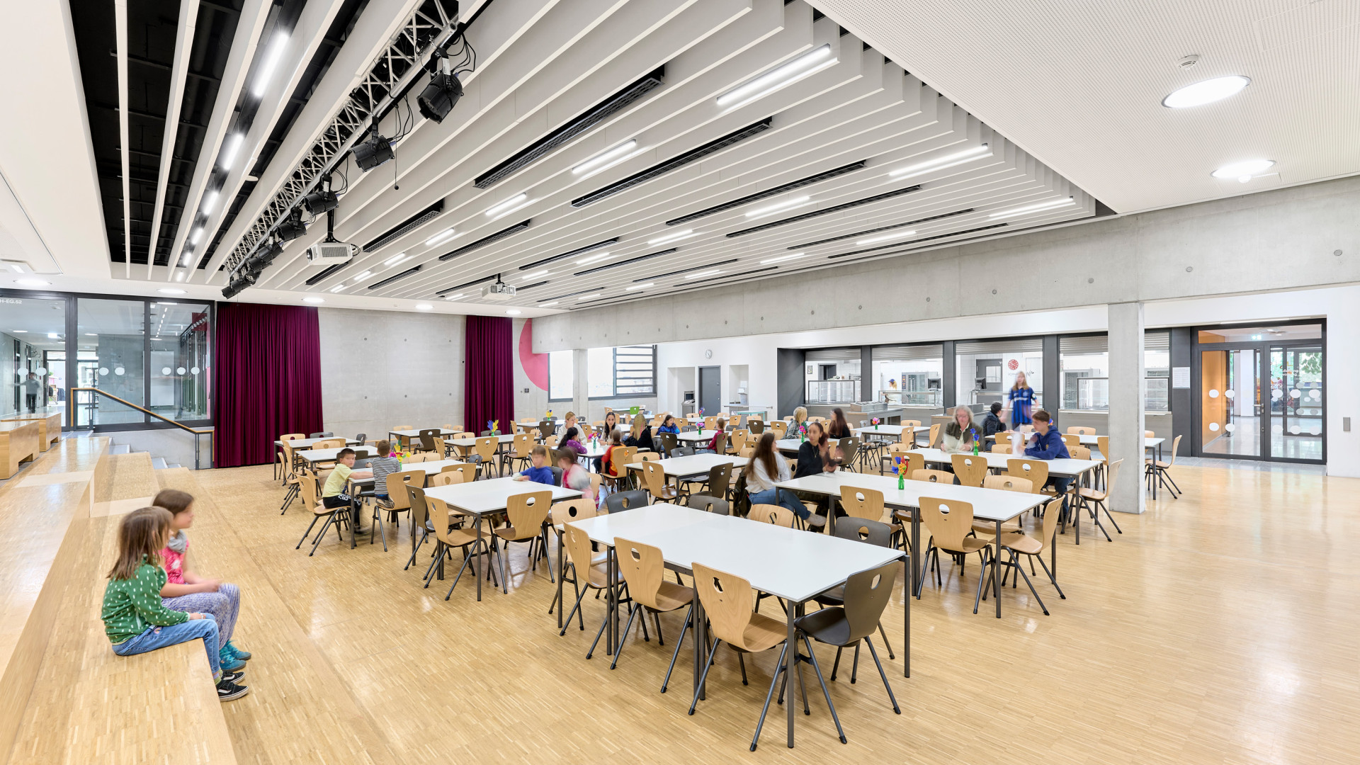 Helle Mensa mit Tischen, Schülern und moderner Architektur im Hintergrund.