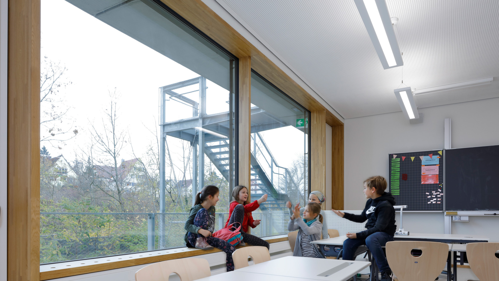 Fünf Kinder in einem modernen Klassenzimmer, spielend am Fenster und angeregt im Gespräch.