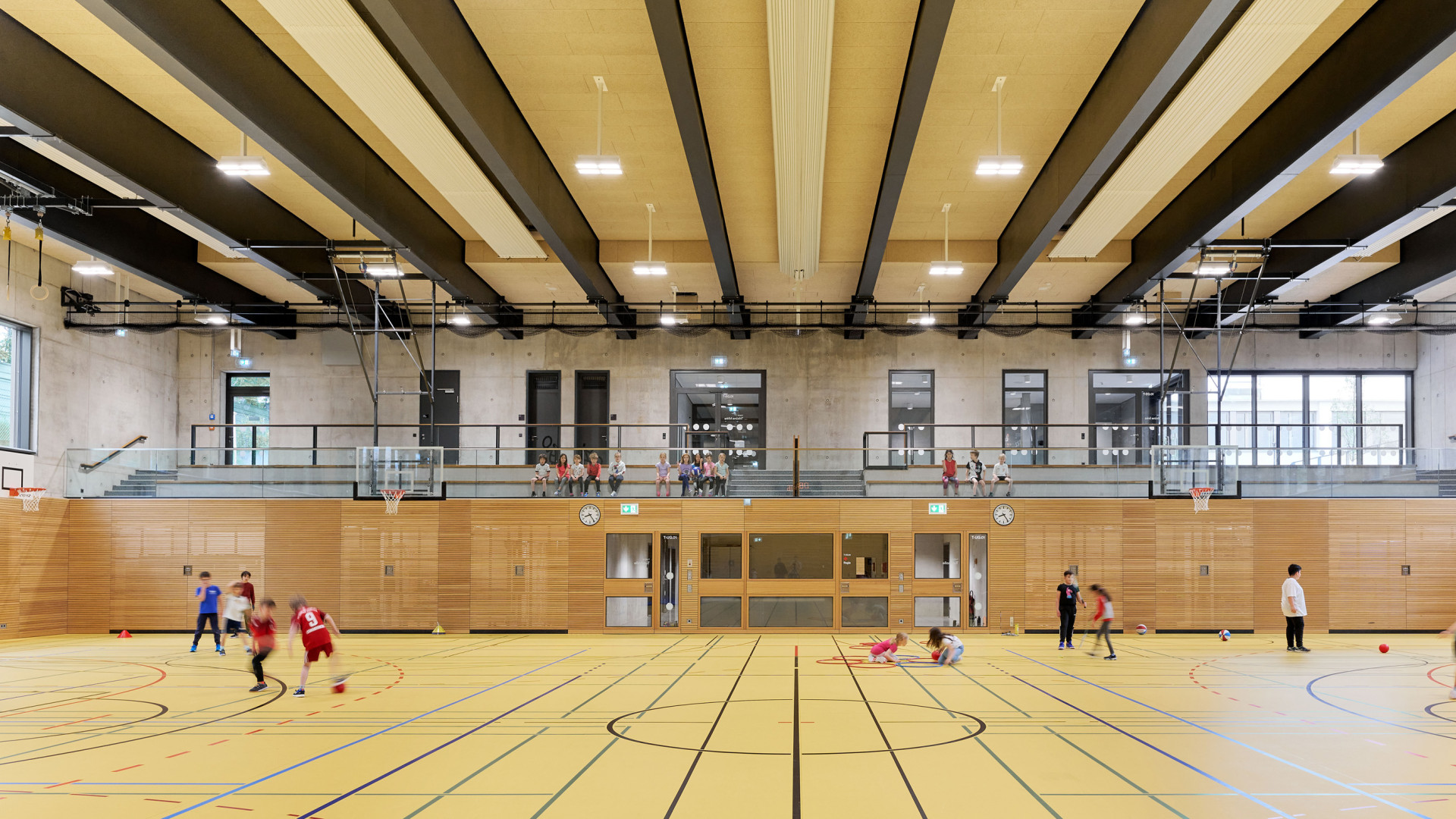 Moderne Sporthalle mit spielenden Kindern und Tribüne im Hintergrund. Helle, ansprechende Atmosphäre.