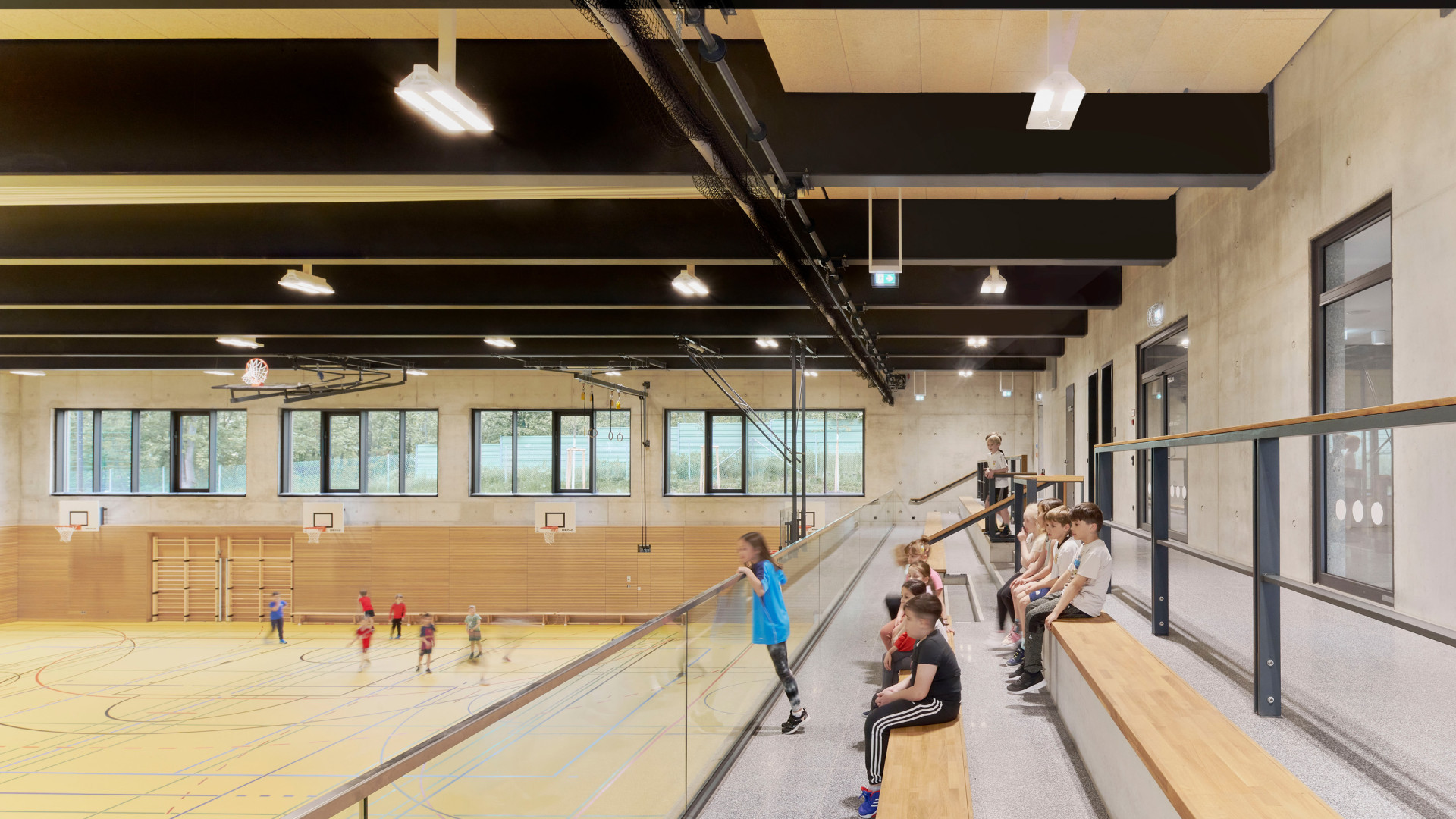 Die lichtdurchflutete Sporthalle zeigt Kinder beim Spielen, Zuschauer auf Holztribünen.