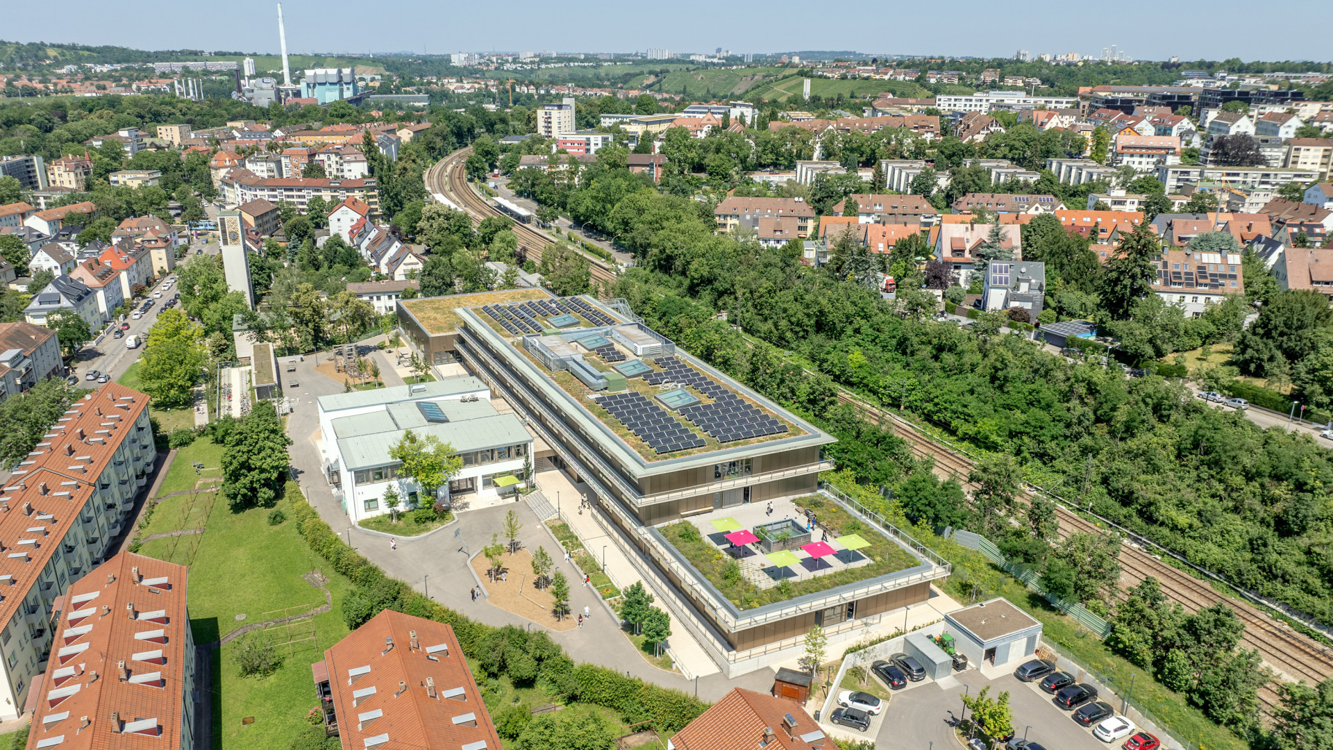 Luftaufnahme eines modernen Gebäudes mit Solaranlagen, umgeben von Wohngebieten und Natur.