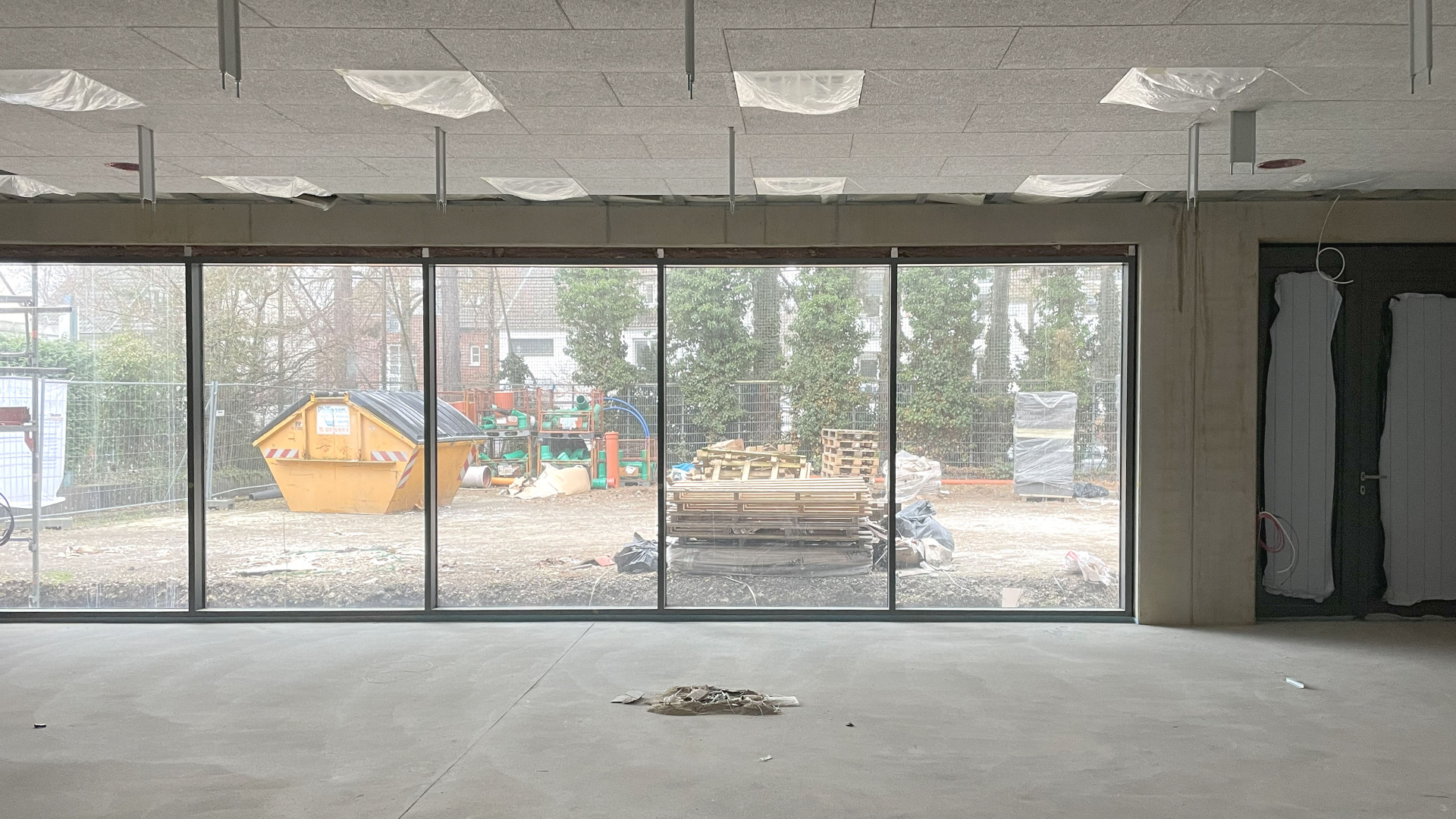 Blick durch große Fenster in einen unvollendeten Raum, Baustellenmaterial und eine gelbe Mulde sichtbar.