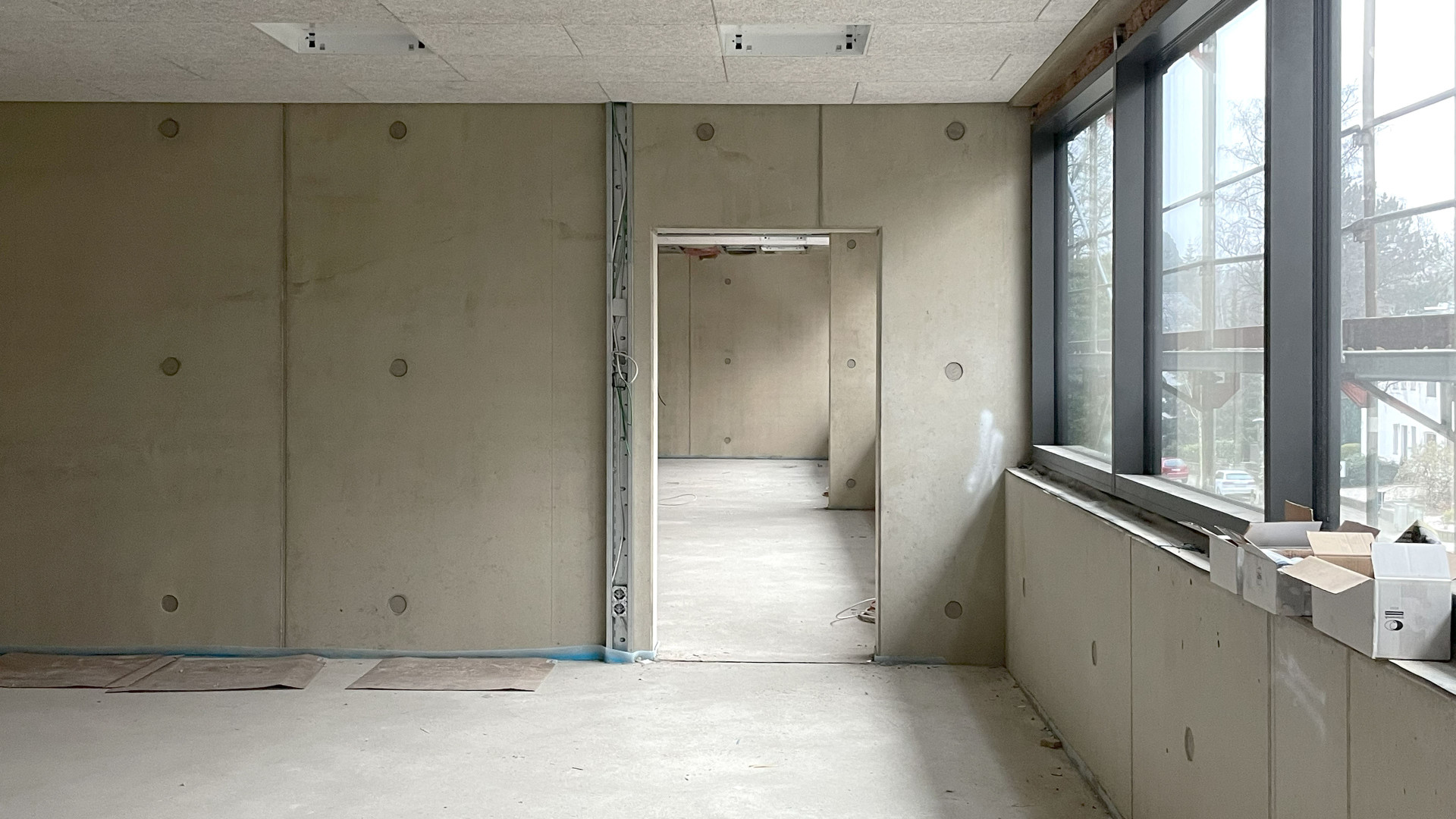 Rohbau eines Raumes mit Betonwänden, Fensterfront und Tür zu einem angrenzenden Raum.