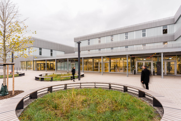 Außenperspektive vom Haupteingang der dreigeschossigen Schule mit runden Sitzbänken auf dem Vorplatz.