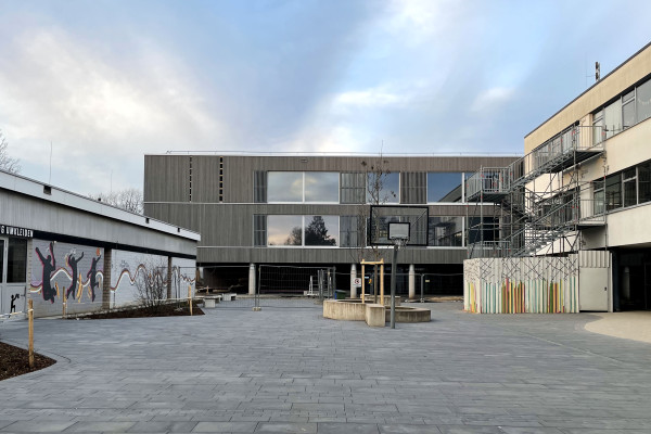 Neues Schulgebäude mit modernem Design, Basketballkorb und Graffiti-Wand auf einem Platz.