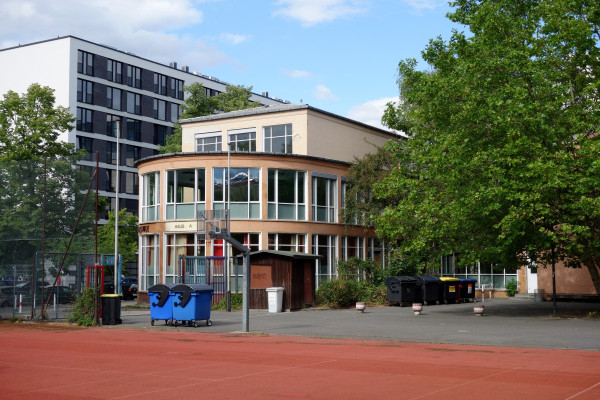 Schule mit großen Fenstern, daneben ein Sportplatz und moderne Wohngebäude im Hintergrund.