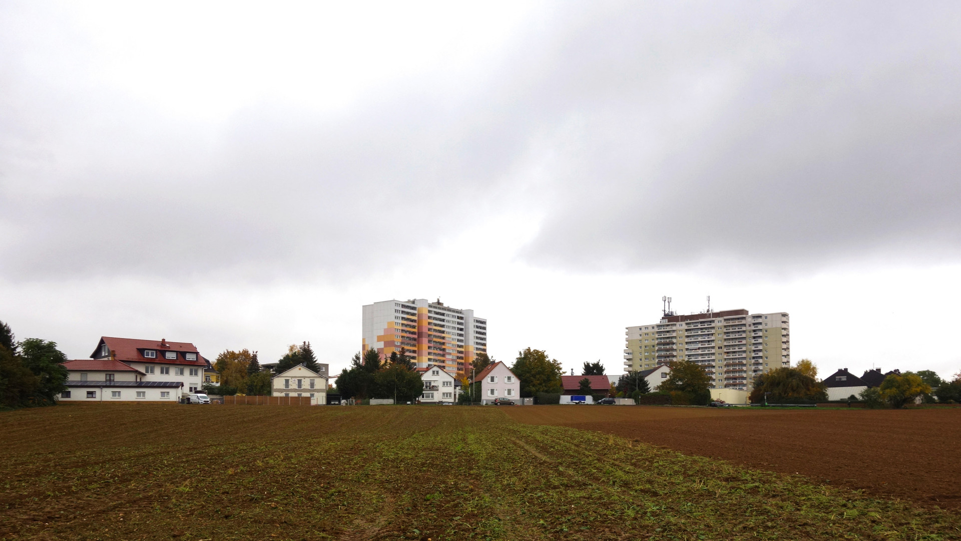 Außenperspektive zeigt zwei Wohnhochhäuser vor denen kleine Einfamilienhäuser und ein großes Feld zu sehen sind.