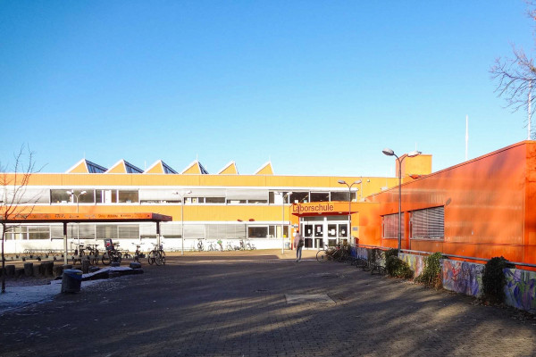 Außenperspektive der zweigeschossigen orangen Laborschule mit der Haupterschließung im Vordergrund.